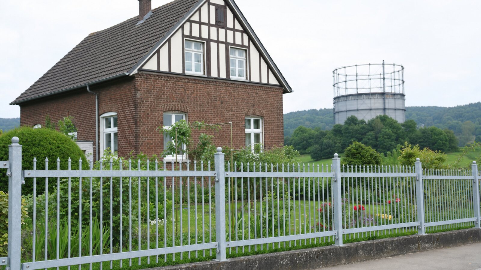 Zaunbau Oberhausen: verzinkter Stabzaun mit Speerspitzen vor einem Backsteinhaus der Arbeitersiedlung, im Hintergrund der Gasometer